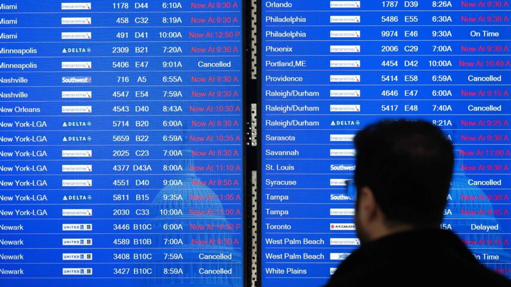 A traveler looks at a large digital airport departures board filled with delayed and cancelled flights, with red text indicating revised departure times, reflecting widespread airline disruptions.