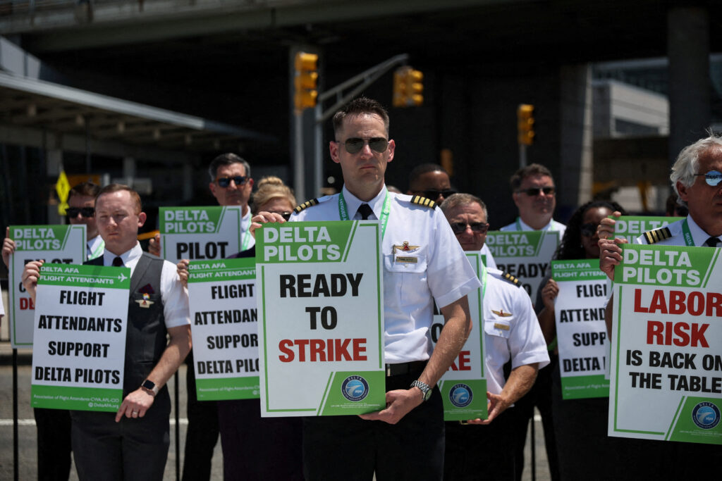 American Airlines pilots in uniform hold signs and protest outside an airport terminal, advocating for better working conditions and pay, amid broader labor negotiations in the airline industry.
