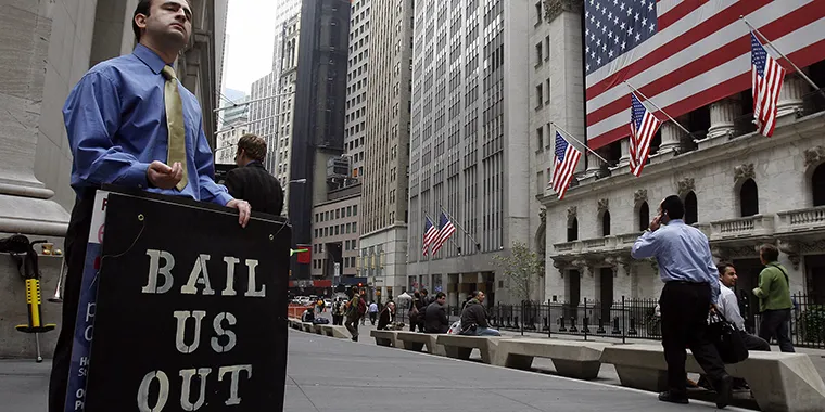 A man in business attire holds a protest sign reading ‘Bail Us Out’ outside the New York Stock Exchange during the 2008 Great Recession.